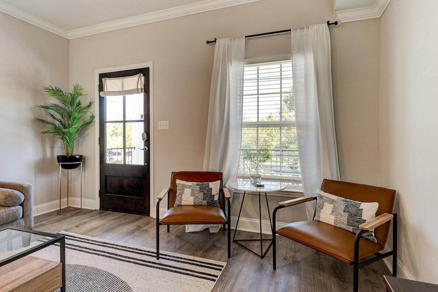 A living room with two leather chairs, a glass coffee table, a potted plant, a door, and a window with curtains.