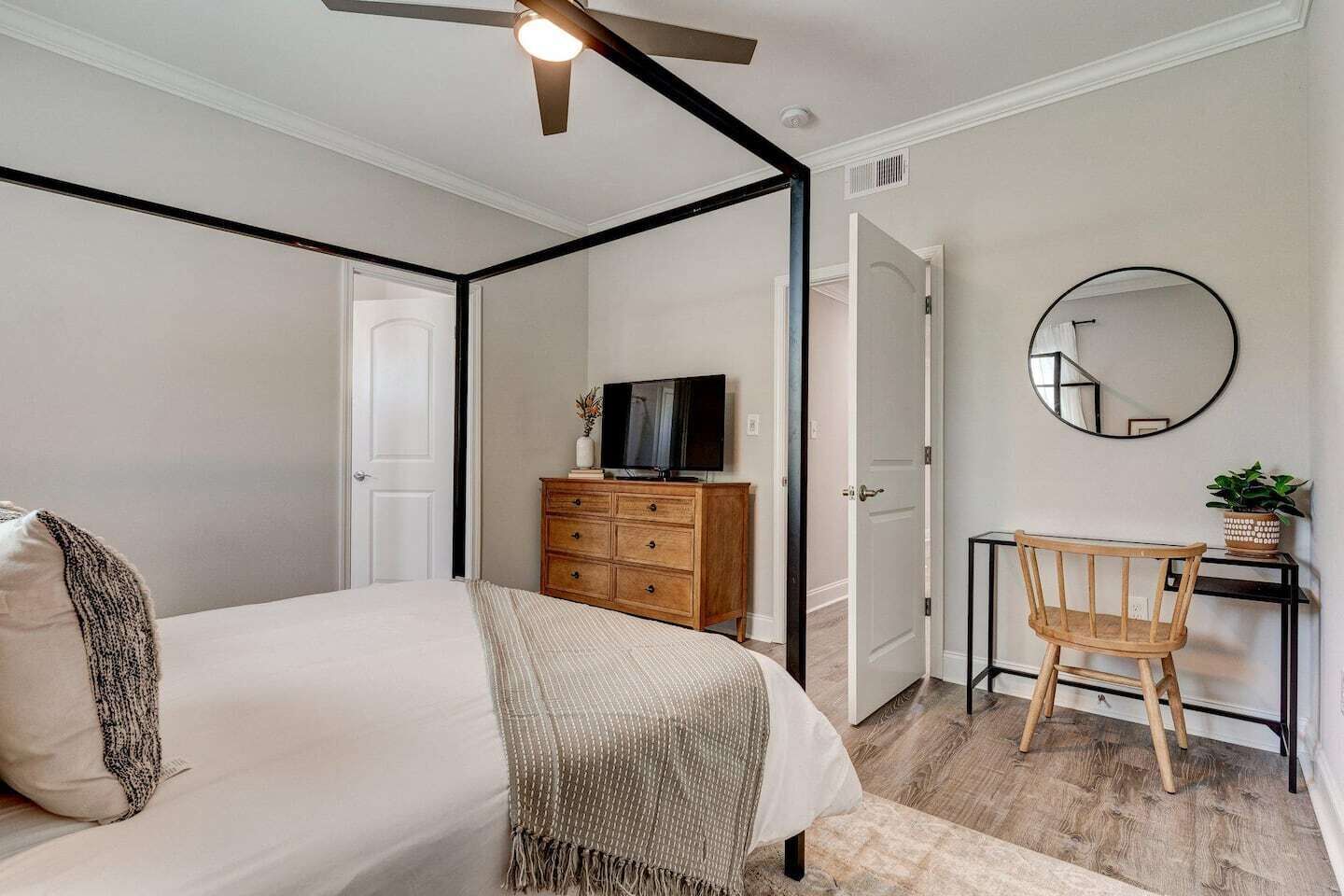 A bedroom with a four-poster bed, a wooden dresser, a television, a desk with a chair, and a round mirror on the wall.