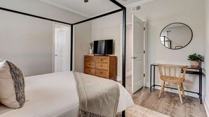A bedroom with a four-poster bed, a wooden dresser, a television, a desk with a chair, and a round mirror on the wall.