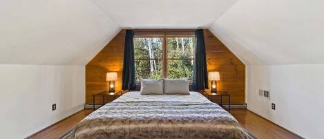 Un dormitorio con techo inclinado, una cama grande, dos lámparas de noche y vistas a árboles a través de una ventana.
