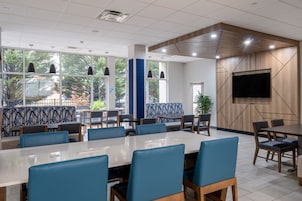 A conference room with a large table and chairs, large windows with a view of trees, and a flat-screen TV mounted on the wall.