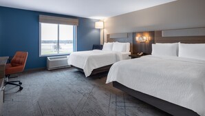A hotel room with two beds, a desk, an orange chair, a large window, and a wall-mounted lamp.