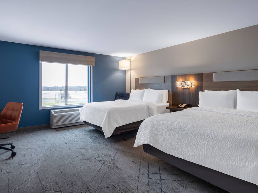 Ein Hotelzimmer mit zwei Betten, einem Schreibtisch, einem orangefarbenen Sessel, einem großen Fenster und einer Wandlampe.