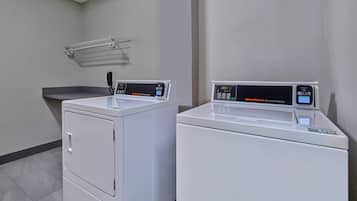 Two commercial washers in a clean, tiled laundry room with a metal drying rack on the wall.