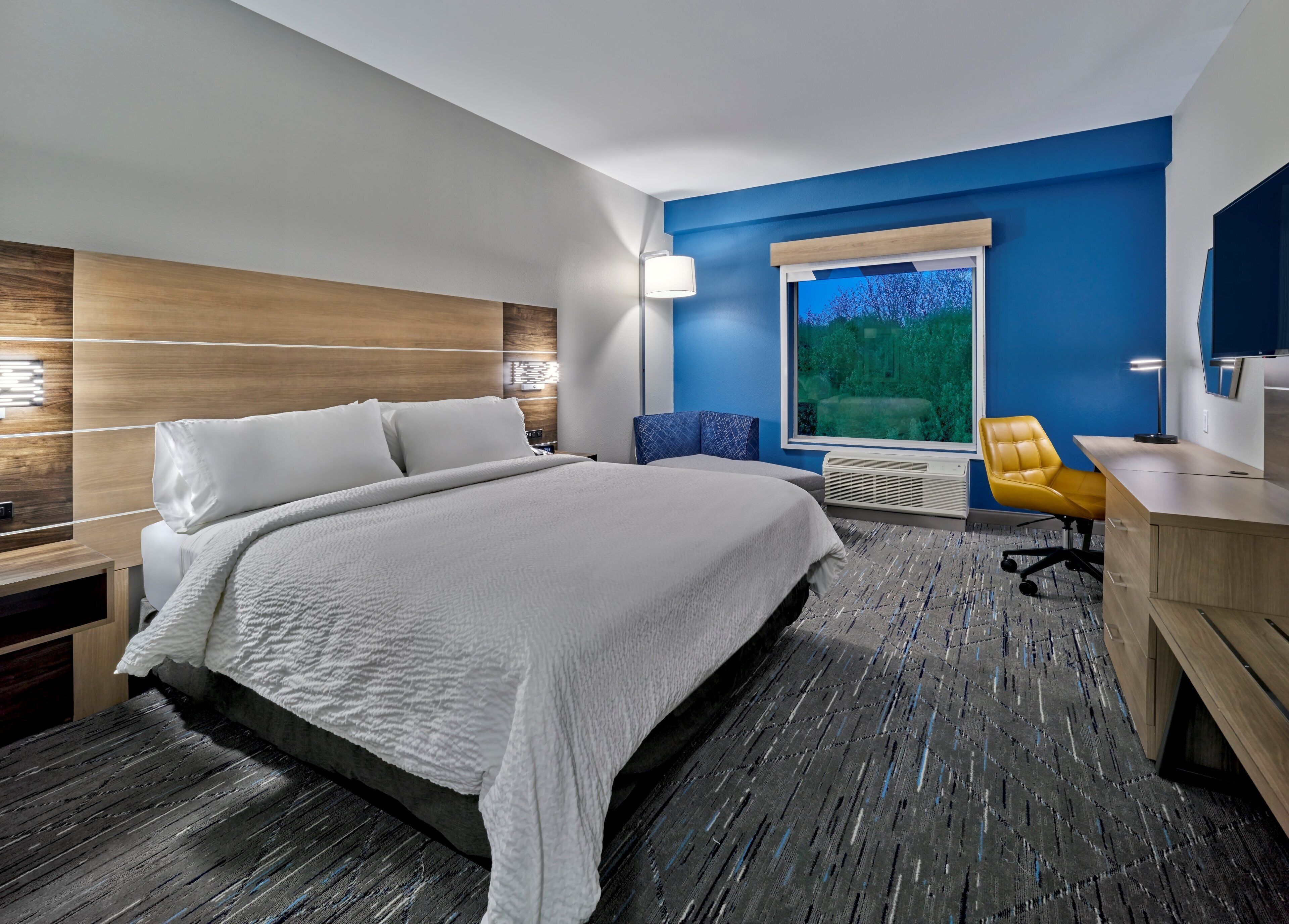 Une chambre d’hôtel moderne, dotée d’un grand lit, d’un bureau, d’une chaise et d’une fenêtre donnant sur la verdure.