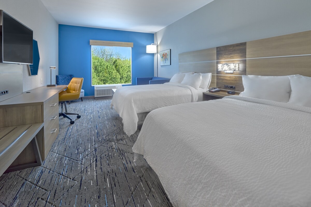Habitación de hotel con dos camas, un escritorio, una silla y un amplio ventanal con vistas a zonas verdes.