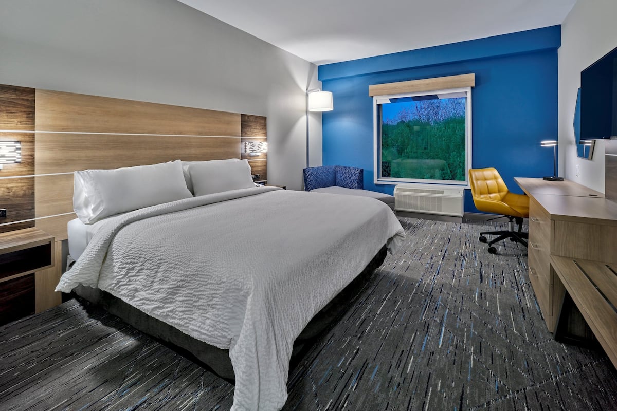 A modern hotel room with a large bed, a desk, a chair, and a window with a view of greenery.