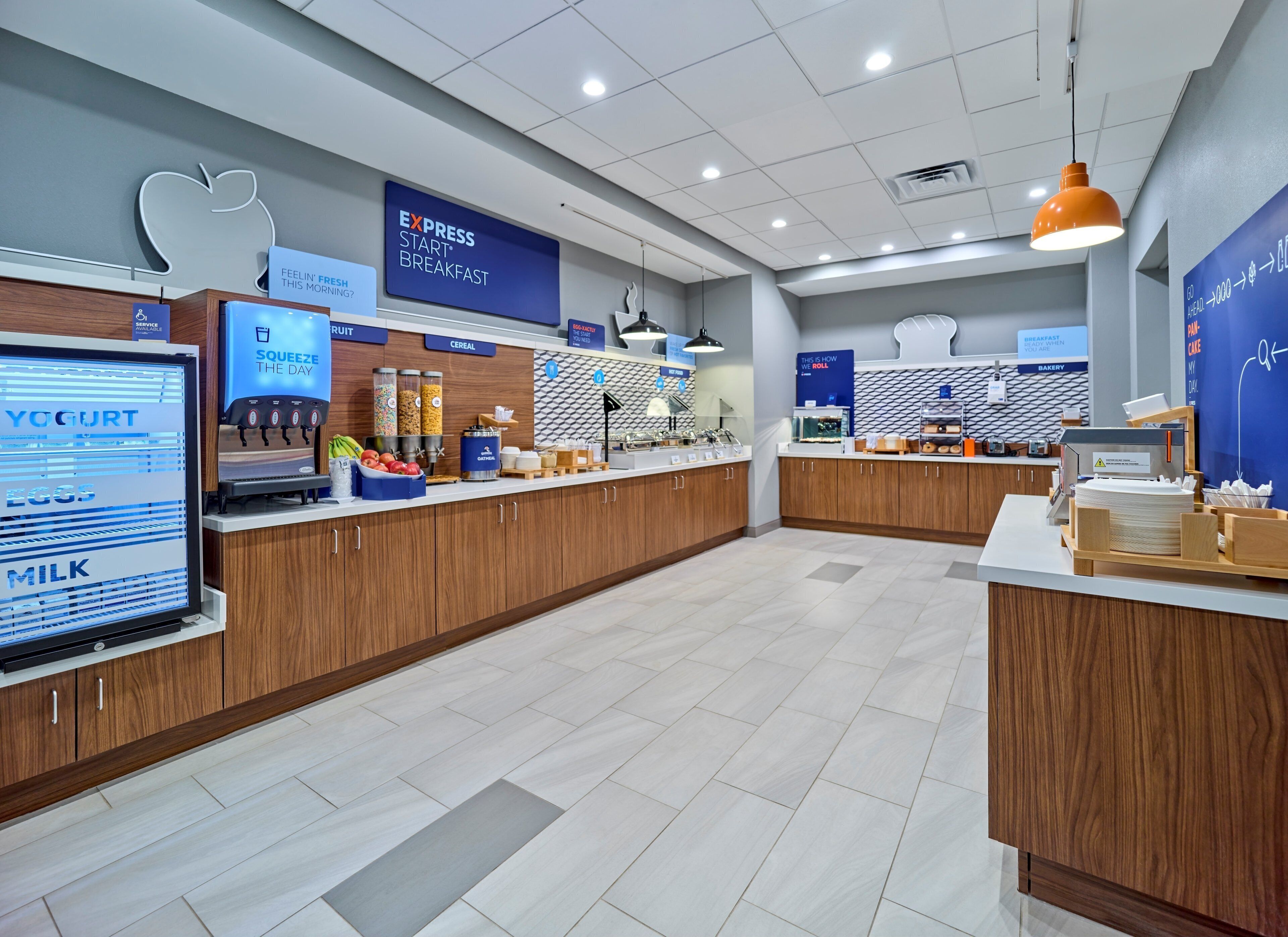 A modern breakfast area with a digital menu board, various food stations including yogurt, cereal, and fruit, and a bakery section.