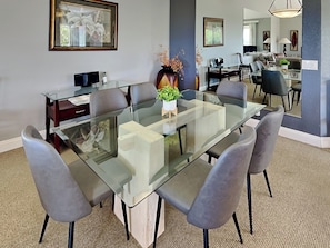 A modern dining room with a glass-top table, grey upholstered chairs, and a vase with flowers. - Grand Champions by Maui Rental Group (Kihei)