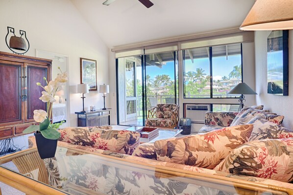 A spacious living room with a large sofa, a dining table, and a view of palm trees through large windows. - Grand Champions by Maui Rental Group (Kihei)