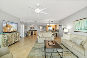 A spacious living room with a large flat-screen TV, a beige sofa, a coffee table, and a ceiling fan.