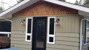 Un petit bâtiment d’un seul étage, avec une porte d’entrée sombre, deux fenêtres de chaque côté et un petit porche.