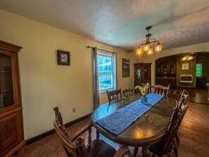 Uma sala de jantar com uma mesa de madeira comprida, cadeiras, um candelabro e uma janela com cortinas.