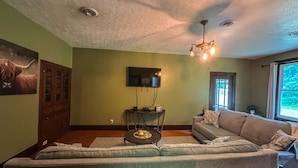 A living room with a sectional sofa, a flat-screen TV mounted on the wall, a ceiling light fixture, and a window with a view of greenery.