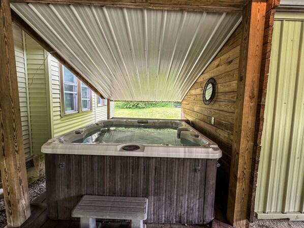 Un jacuzzi con un banco de madera frente a él, ubicado en un espacio exterior cubierto con paredes de madera y vistas a un paisaje verde.