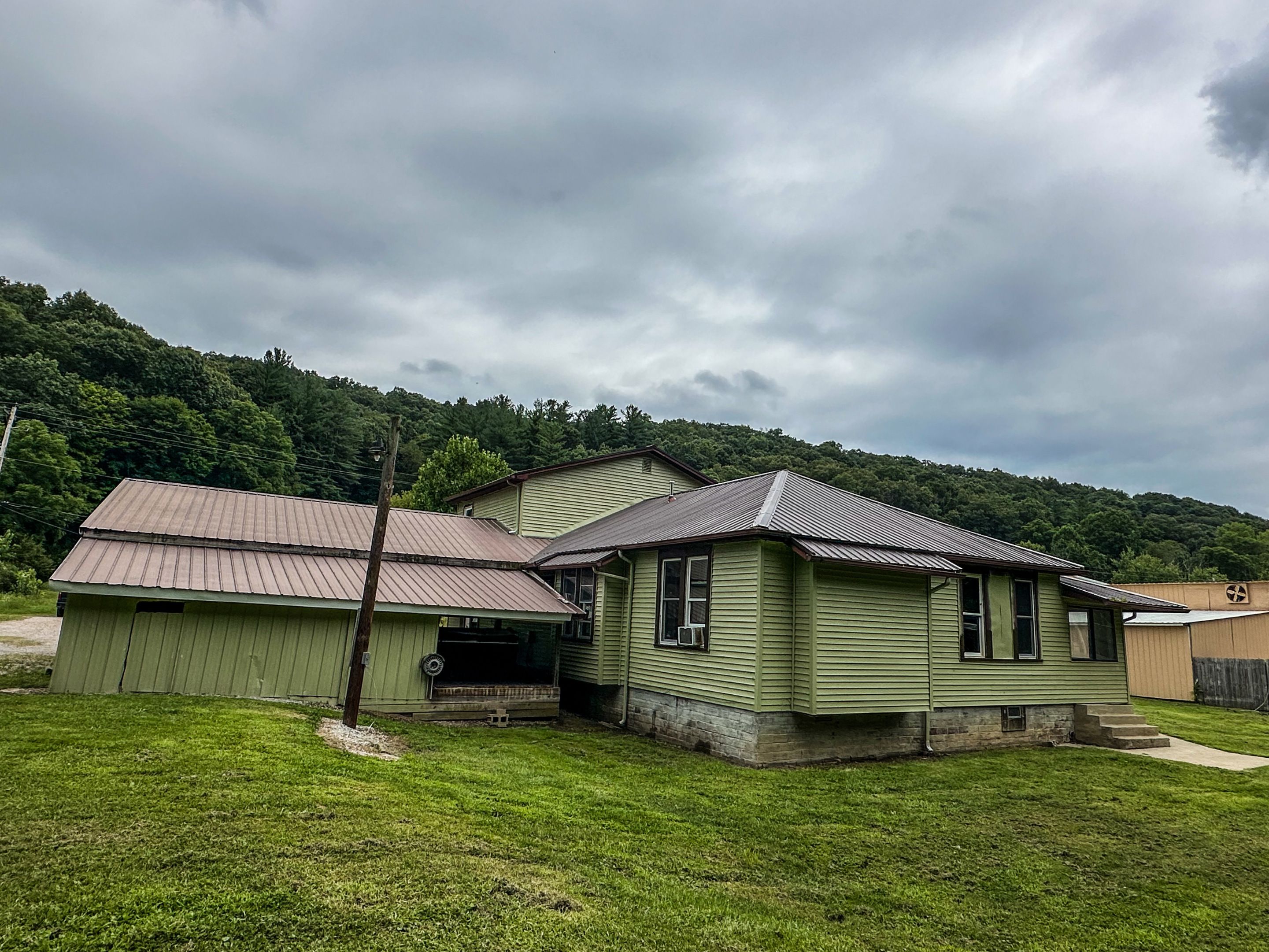 A two-story house with a green exterior and a metal roof, surrounded by a grassy yard and a forested hill in the background.