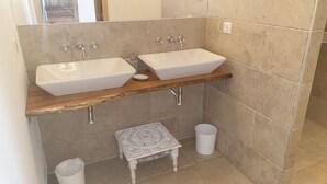 Une salle de bain avec deux vasques sur pied, une étagère en bois, une petite table décorative et une poubelle.