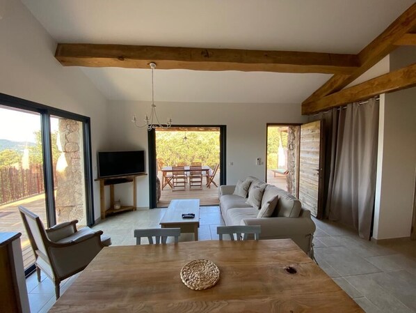 A spacious living area with a wooden dining table, a sofa, and a television. There are large windows and a view of the outdoors.