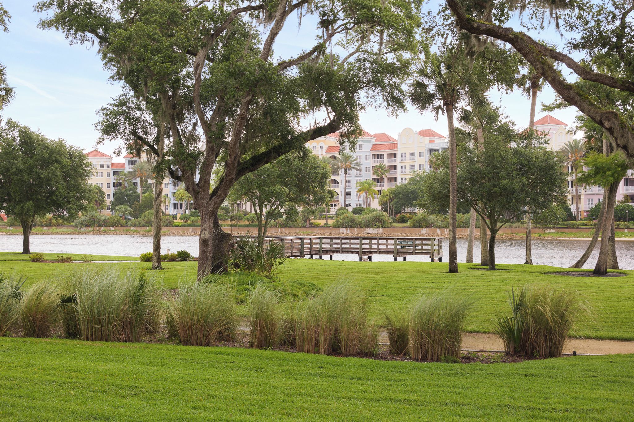 A park with a lake, trees, and a wooden bridge. In the background, there are buildings with red roofs.