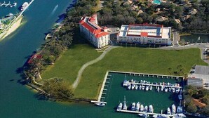 Vista aèria d'un port esportiu amb embarcacions amarrades, un edifici gran amb teulada vermella i una zona verda.