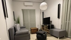 A modern living room with a grey sofa, a small ottoman, a wooden side table, a floor lamp, and a flat-screen TV. - Enjoy Chania (Chania)