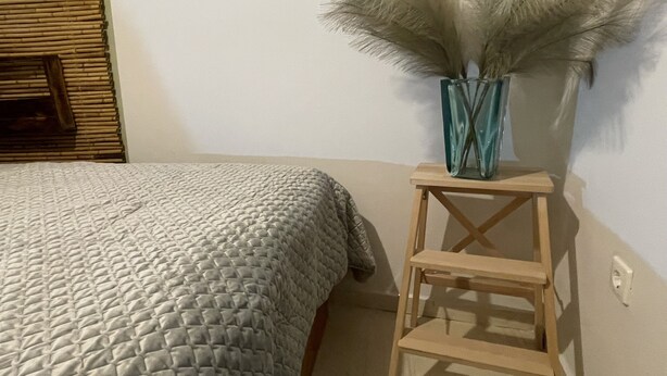 A bed with a textured blanket, a small wooden stool with a vase of pampas grass, and a wall with a switchboard. - Enjoy Chania (Chania)