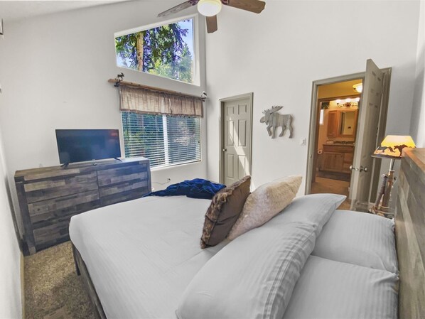 Une chambre à coucher comprenant un lit, une commode, un téléviseur, une fenêtre avec des stores, un ventilateur de plafond et une porte donnant sur une autre pièce.