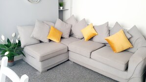 A modern living room with a grey sectional sofa, two yellow cushions, a grey carpet, and a wall-mounted clock. - Swansea Beach Townhouse (Swansea)