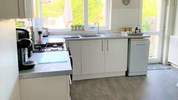 A kitchen with white cabinets, a stove, a sink, and a dishwasher. There are windows with plants on the windowsill and a door with a mat. - Swansea Beach Townhouse (Swansea)