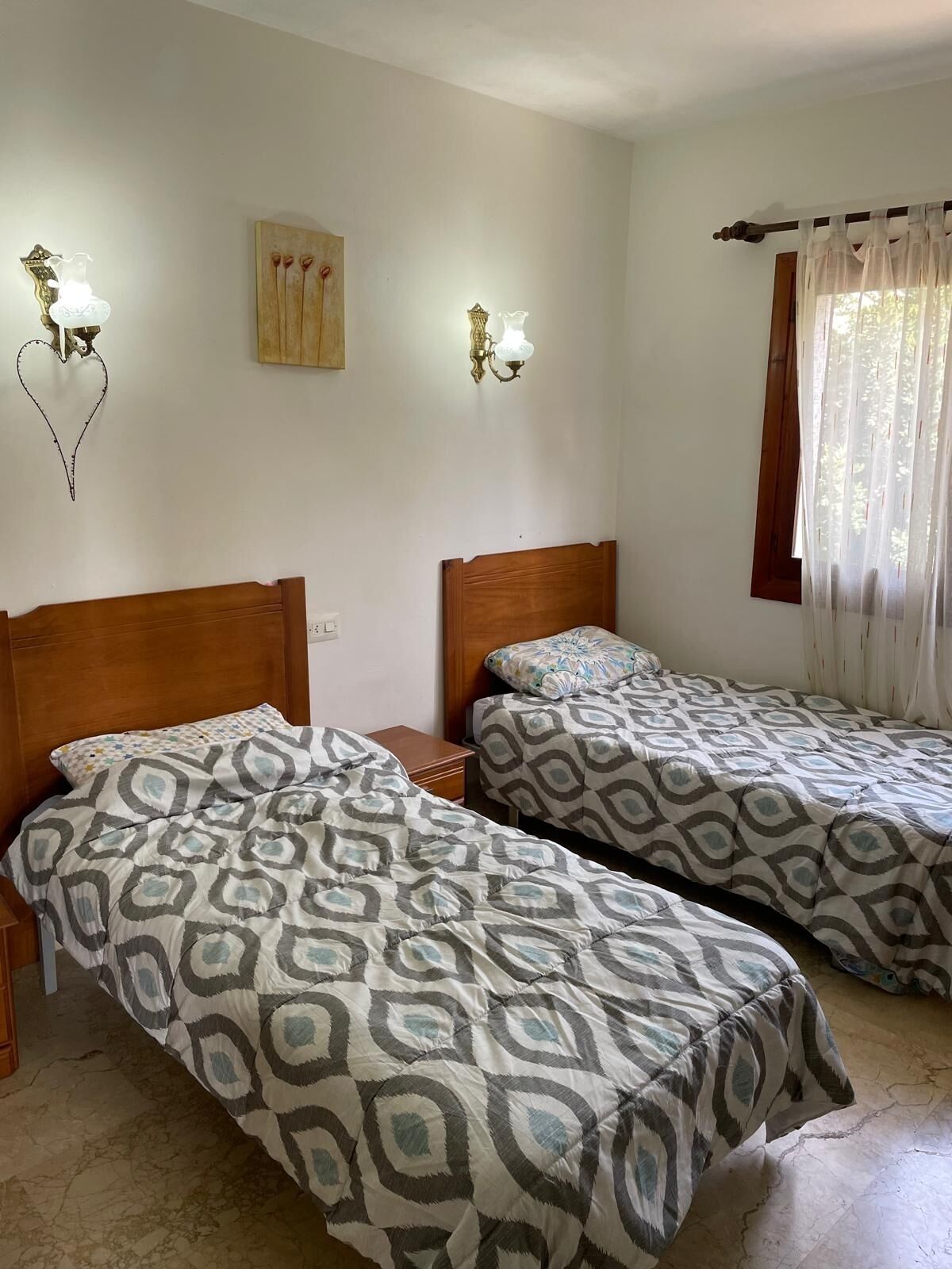 A room with two beds, each with patterned bedding, a wooden headboard, and wall-mounted light fixtures.