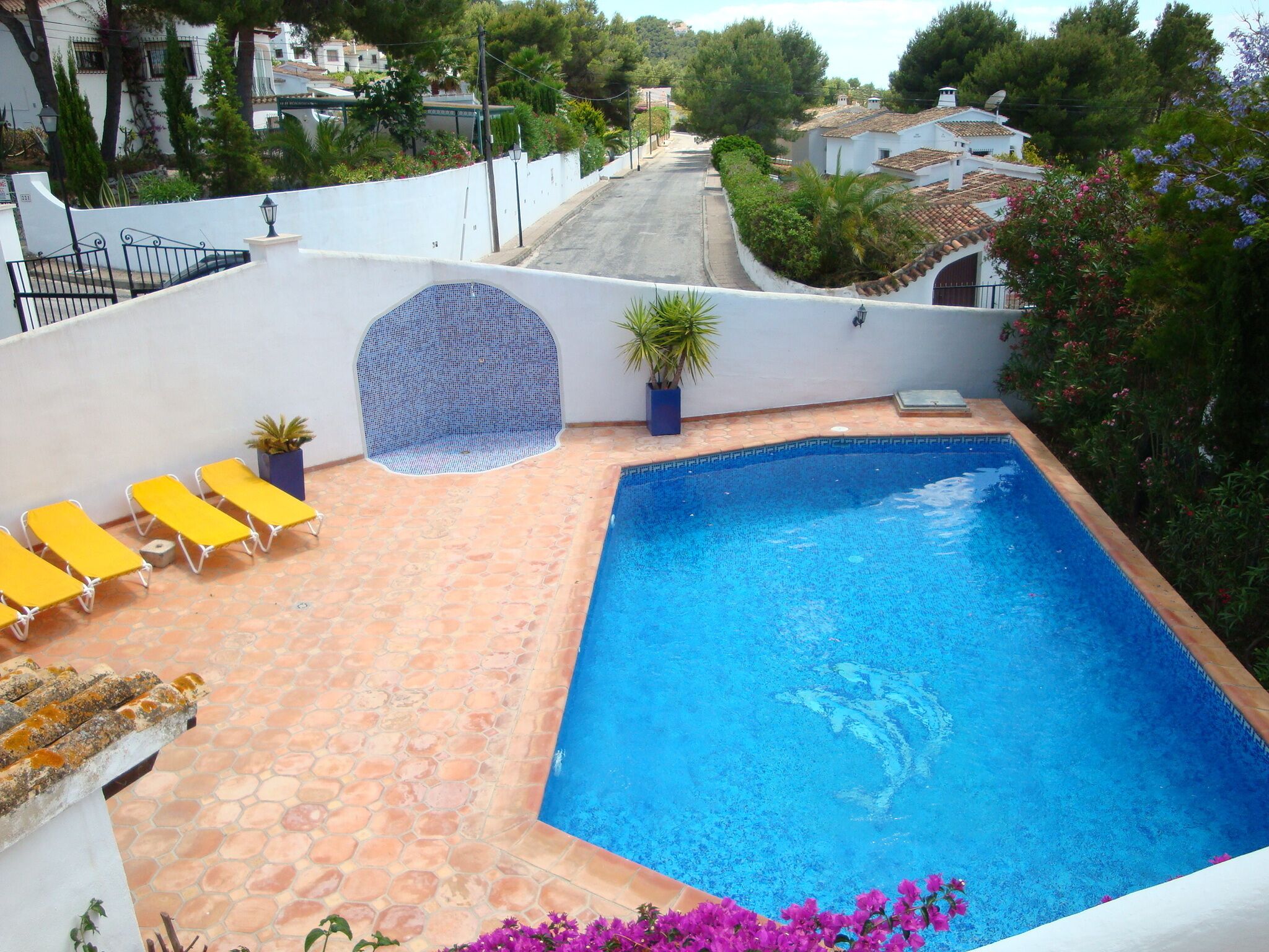 A swimming pool with a tiled deck, surrounded by a white wall and a garden with pink flowers.