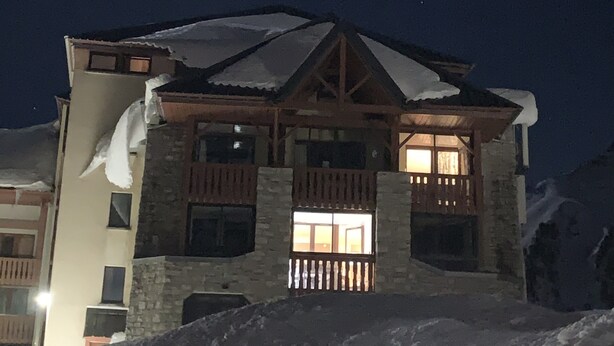 A snow-covered lodge with a balcony and a lit window, under a night sky with a visible moon.