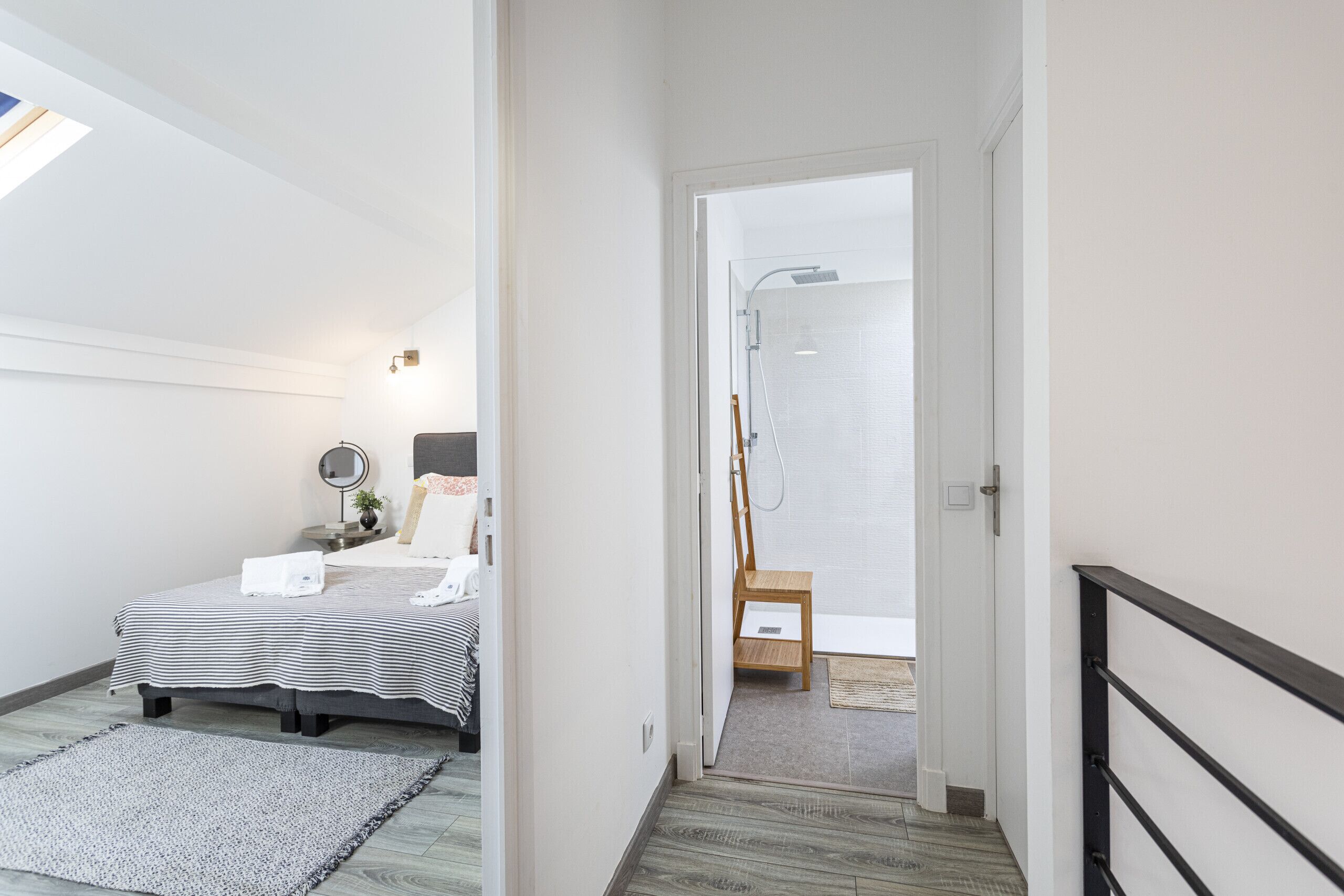 Ein Schlafzimmer mit Bett, Nachttisch und einem kleinen Hocker. Durch eine offene Tür ist ein Badezimmer zu sehen, das eine Dusche und einen Handtuchhalter beinhaltet.