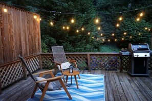 A wooden deck with string lights, two chairs, a small table, and a grill.