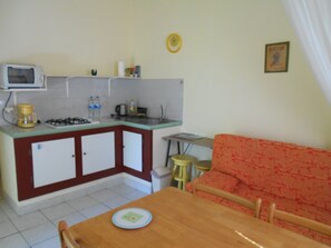 Een compacte keuken met een magnetron, gasfornuis en een klein spoelgedeelte. Er is een eettafel met stoelen en een rode bank in de woonkamer.