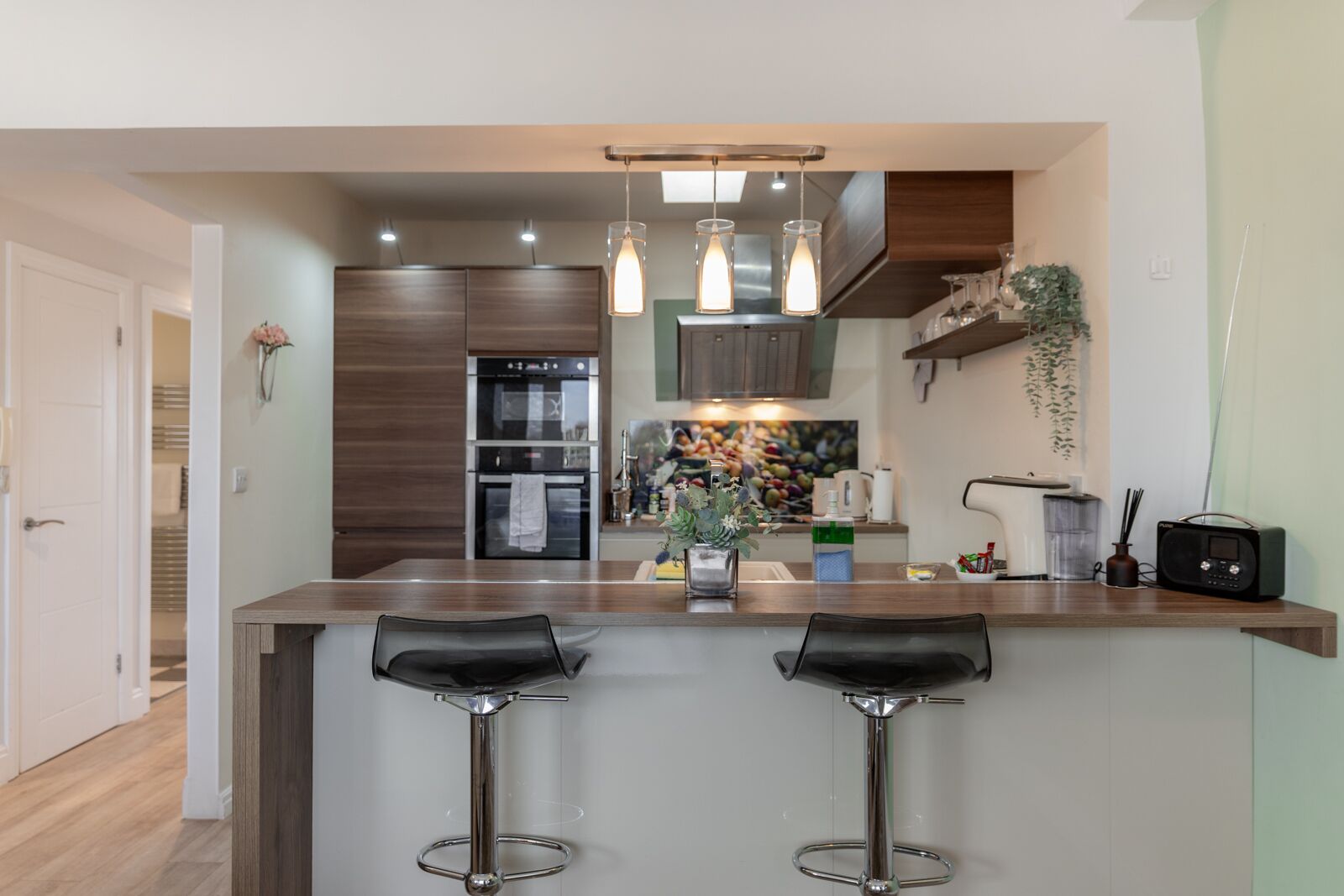 A modern kitchen with two bar stools, a built-in oven, a refrigerator, and pendant lighting.