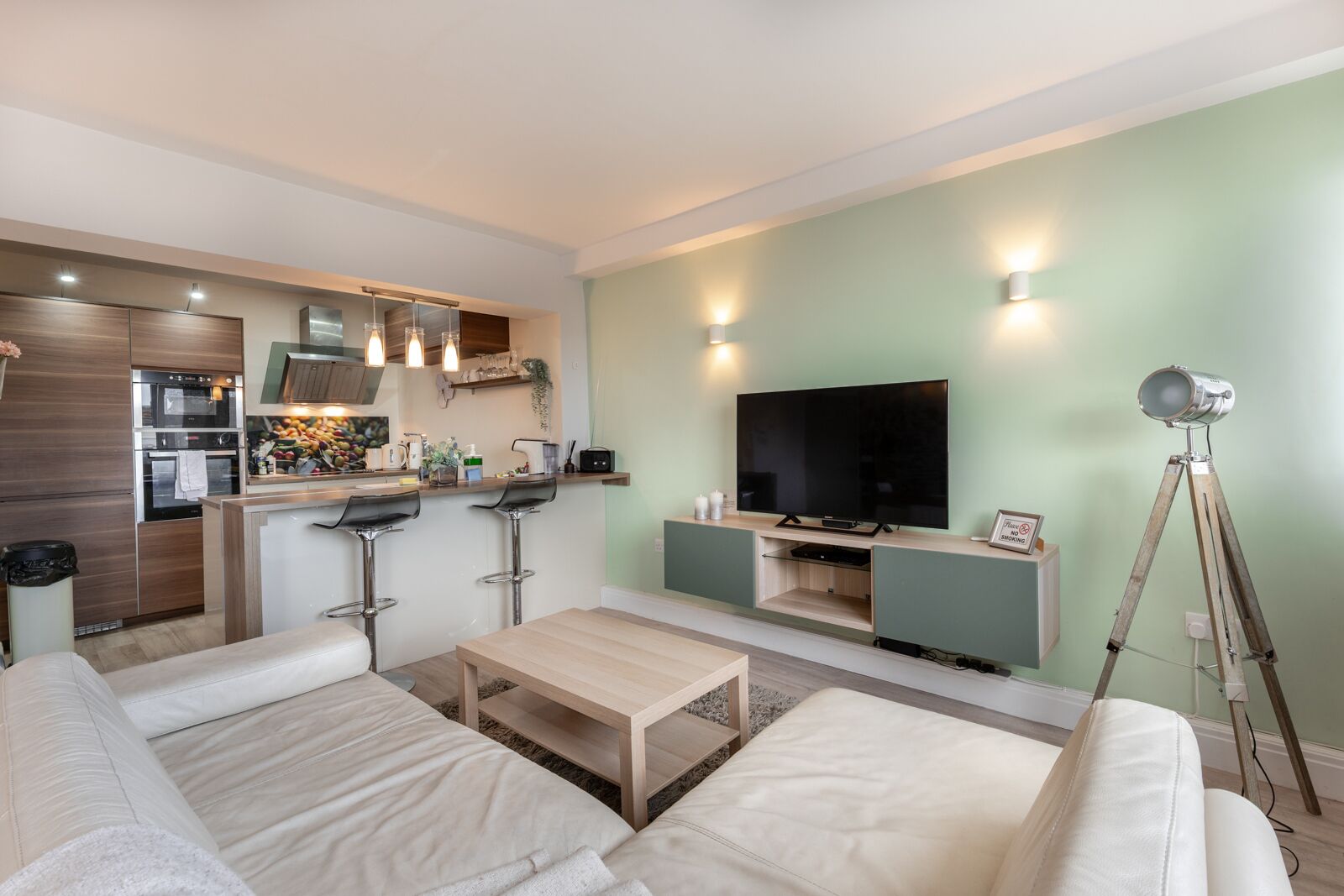 A modern living room with a sofa, a coffee table, a TV, and a kitchen area with a fridge, oven, and bar stools.
