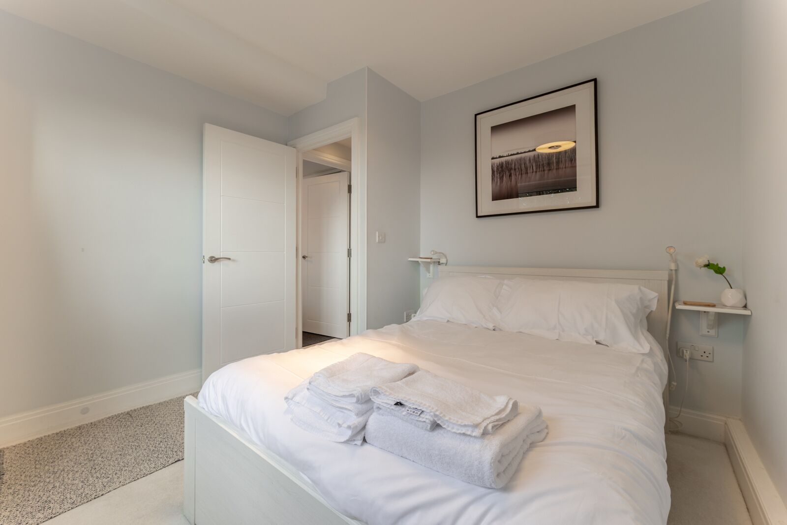 A bedroom with a bed, a door, a framed picture, a nightstand, and a small shelf.