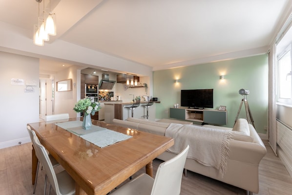 A modern living room with a wooden dining table, a sofa, a TV, and a kitchen in the background.