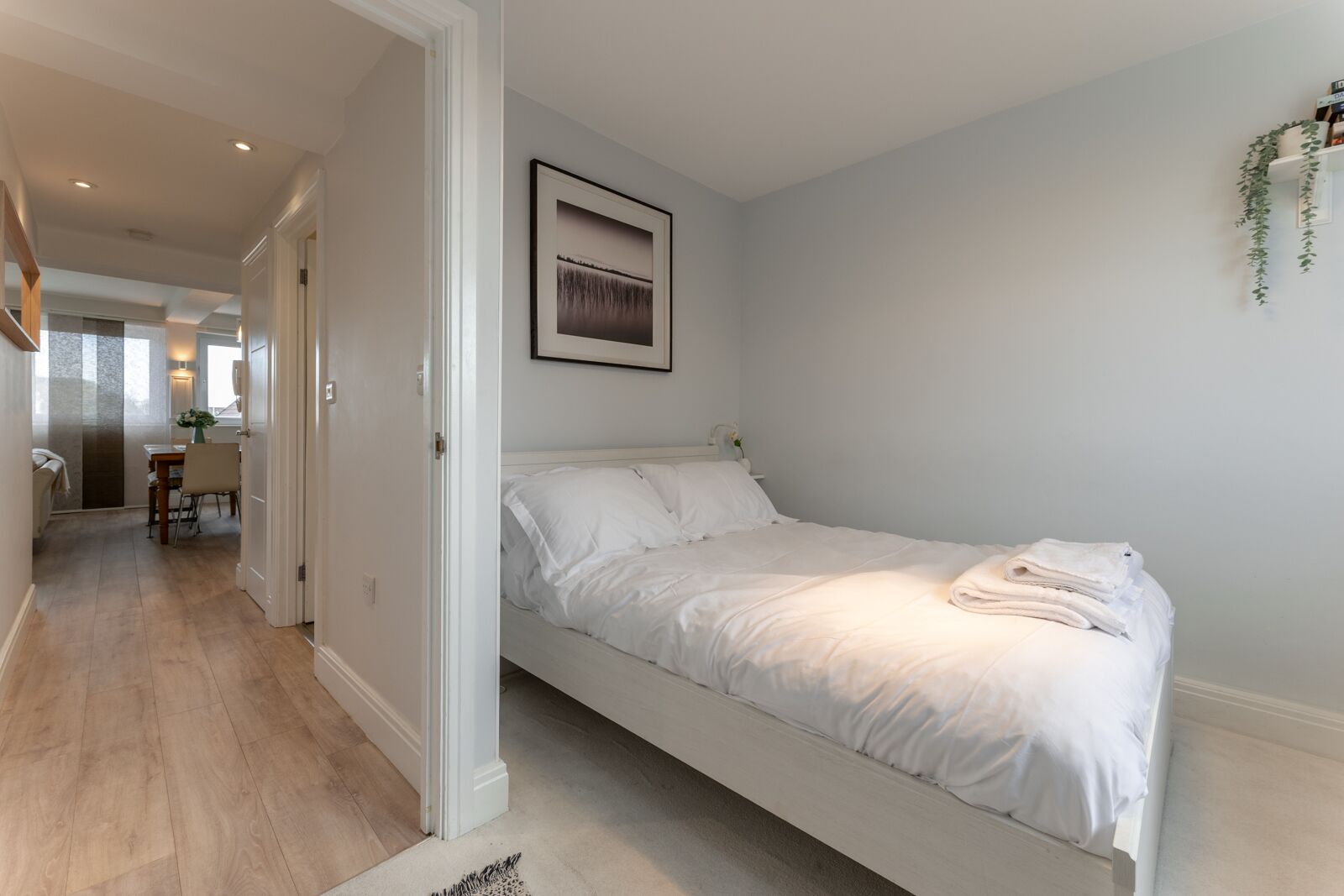 A bedroom with a bed, a framed picture on the wall, and a plant on the right.