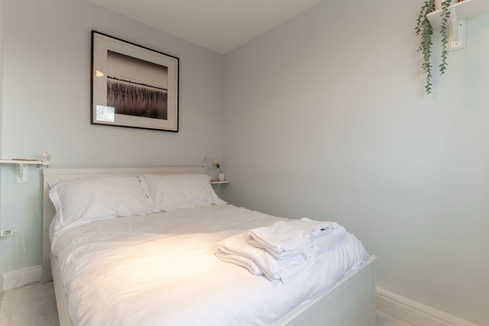 A neatly made bed with white linens, a bedside table with a lamp, and a framed picture on the wall.