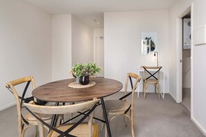Une table ronde en bois, surmontée d’un set de table en fibres tressées, est entourée de quatre chaises en bois dont les pieds sont en métal noir. Un pot de fleurs avec un feuillage vert est placé sur la table.