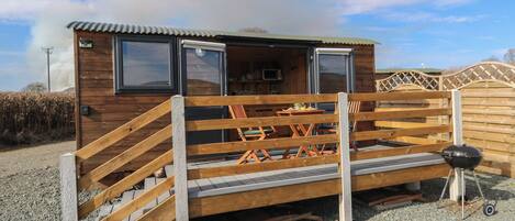 Eine Holzblockhütte mit einer Rampe zu einer Terrasse, einem Grill und einer kleinen Küchenzeile inklusive Mikrowelle.