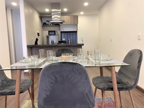 A modern dining area with a glass table and dark chairs, a kitchen in the background, and a wooden floor.
