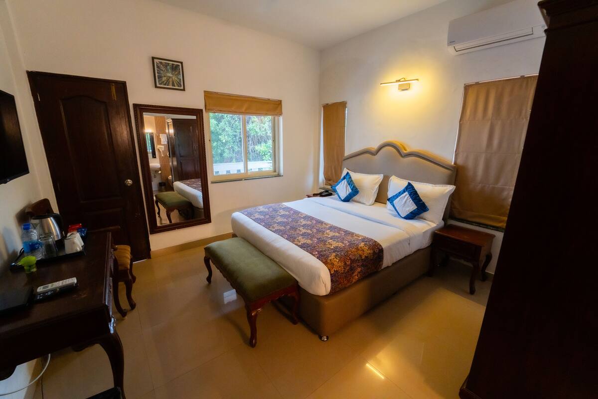 A hotel room with a large bed, a desk with a chair, a small table, and a mirror.