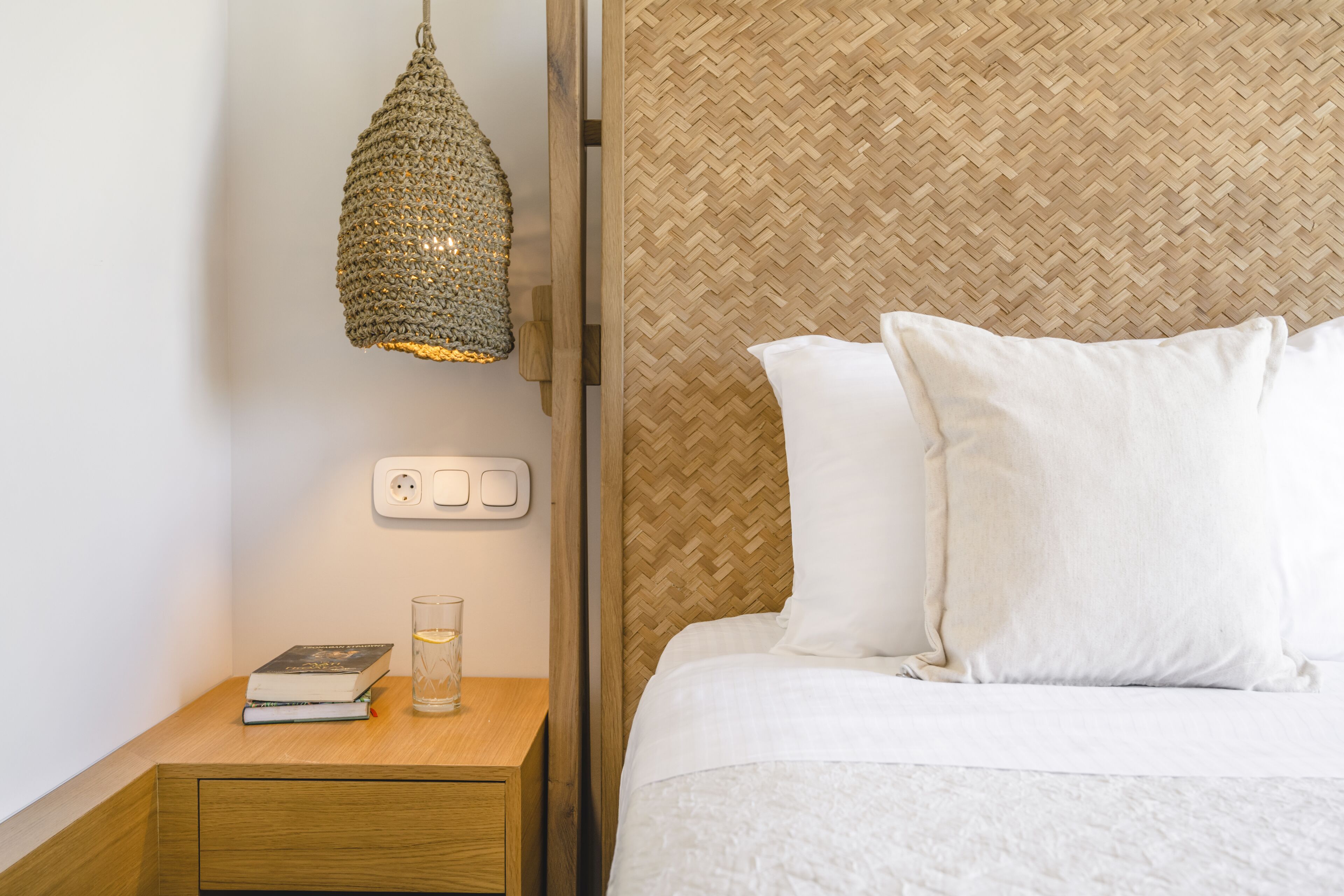 A neatly made bed with white linens, a bedside table with a book and a glass, and a woven wall panel.