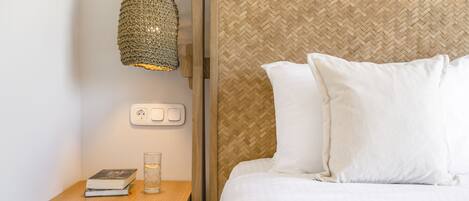 A neatly made bed with white linens, a bedside table with a book and a glass, and a woven wall panel.