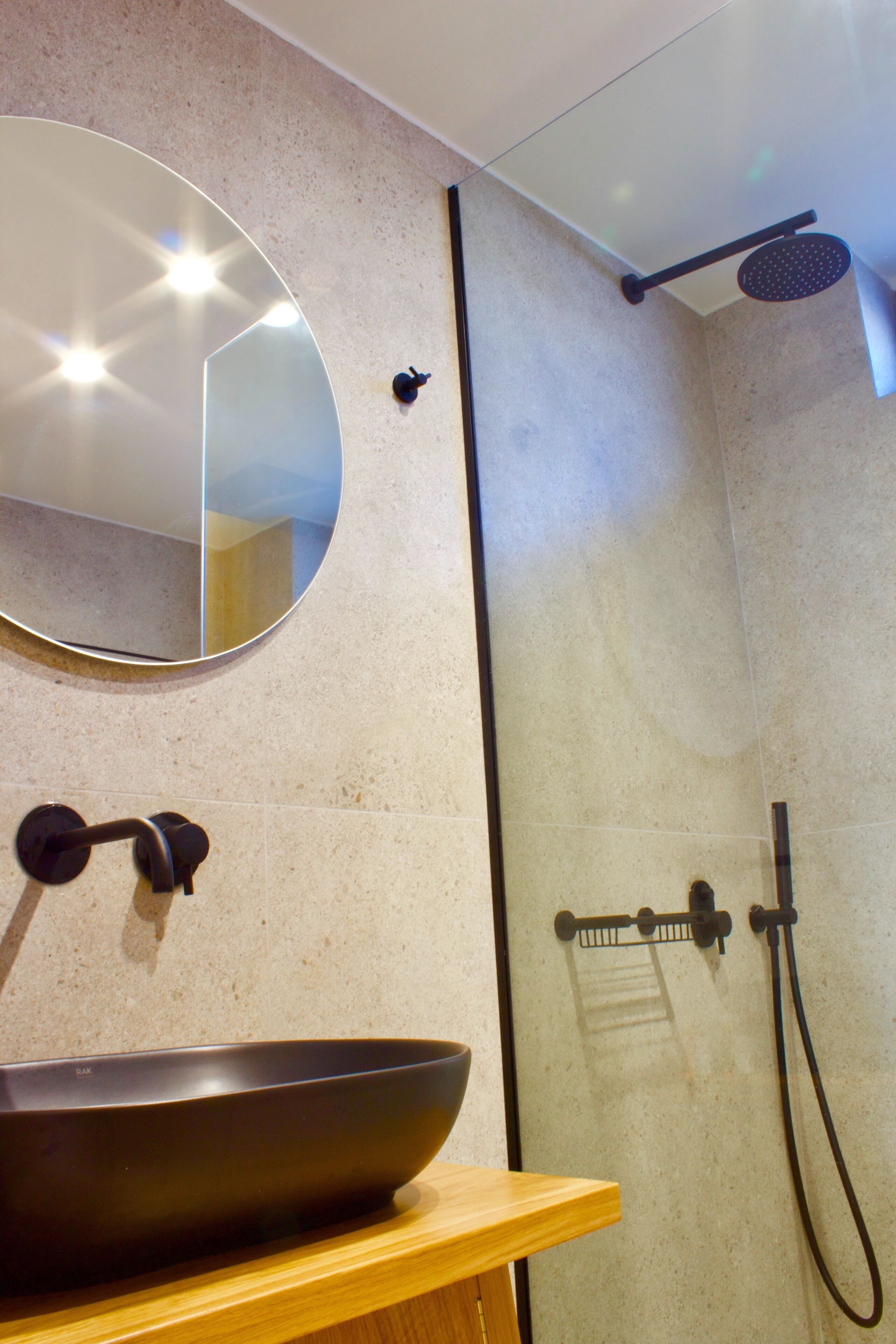 A bathroom with a round mirror, a wooden countertop, a black basin, a glass shower enclosure, and a handheld showerhead.