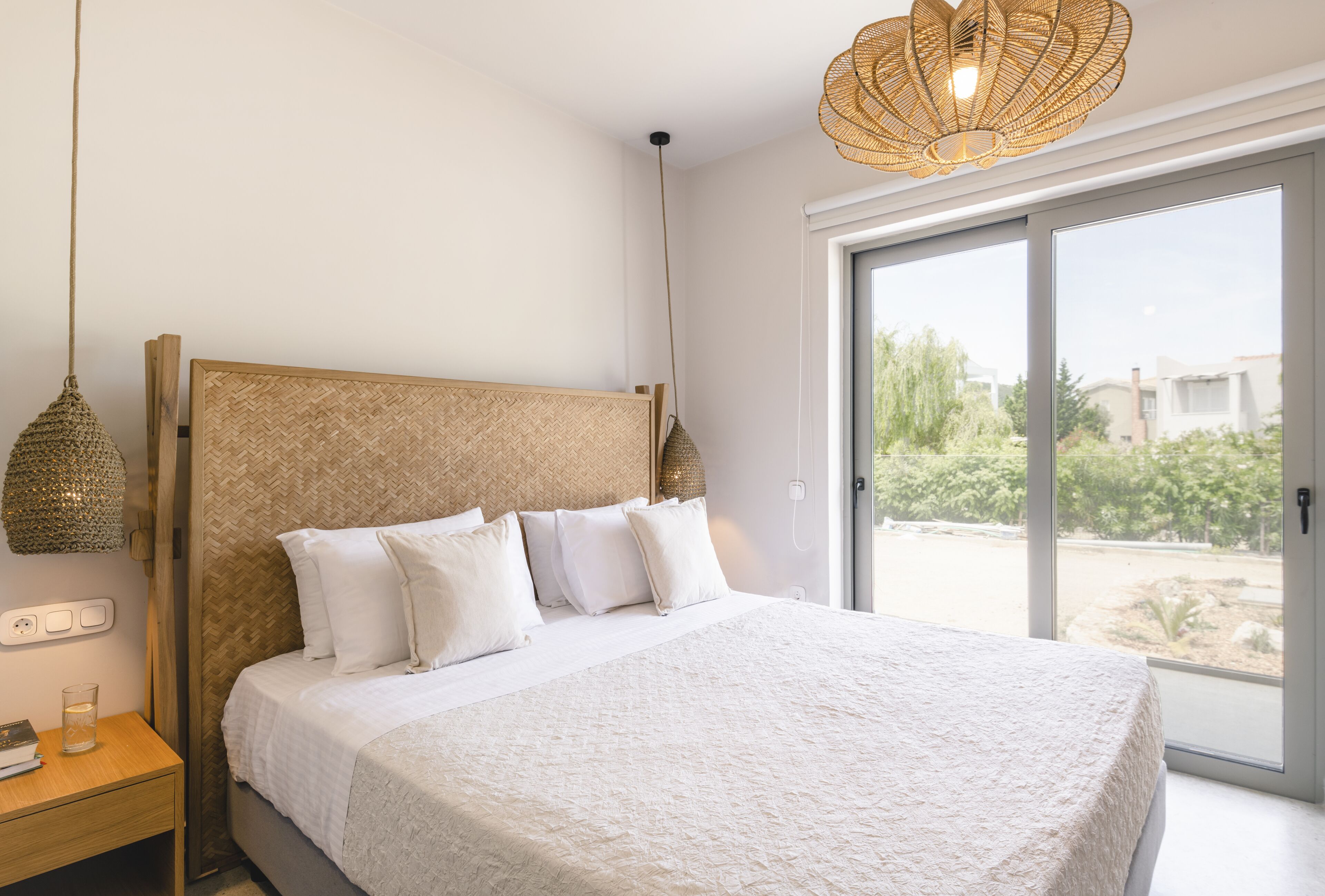 A bedroom with a large bed, a nightstand, a wicker lamp, and a view through a sliding glass door.