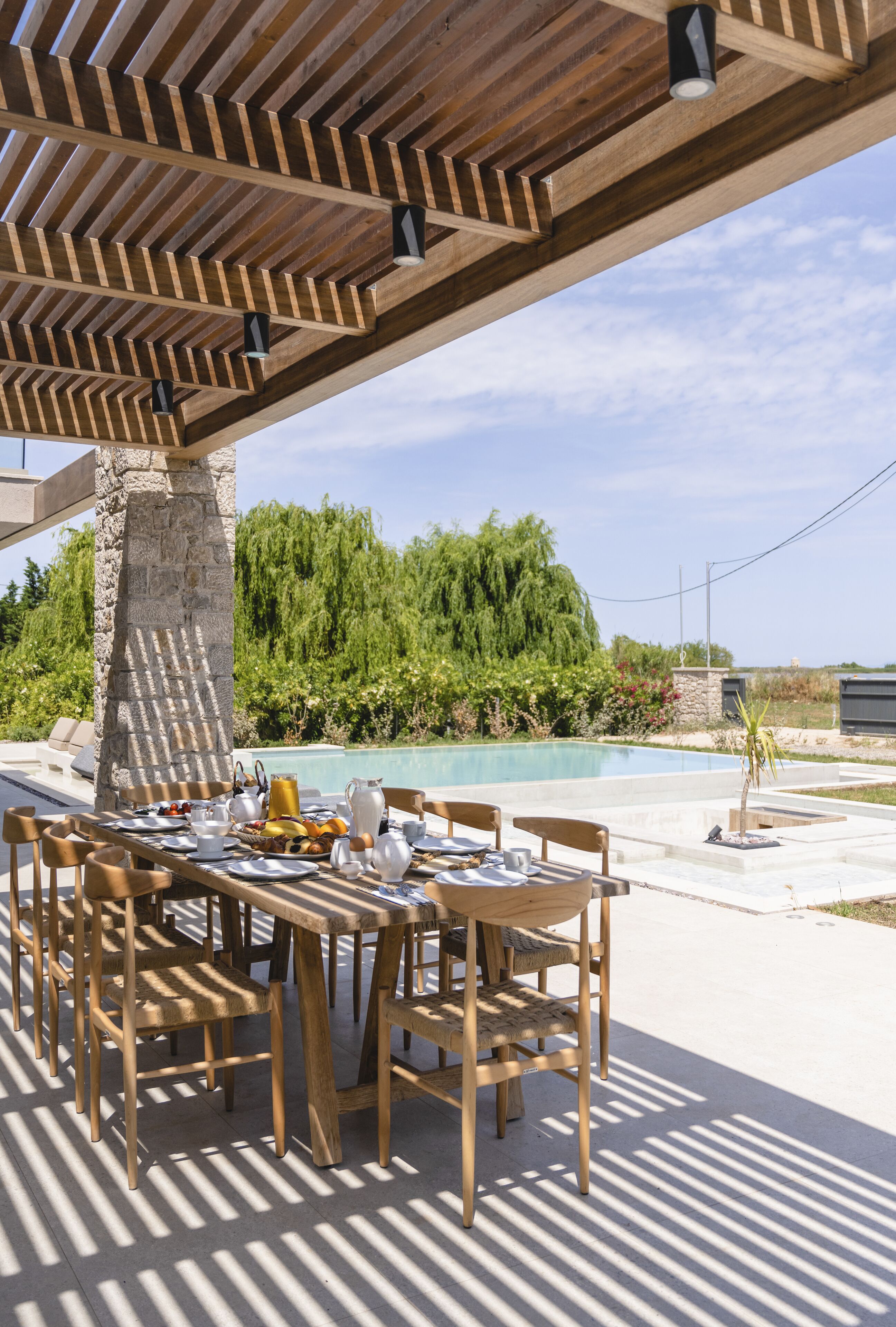 An outdoor dining area with a wooden pergola, a stone pillar, and a pool in the background.